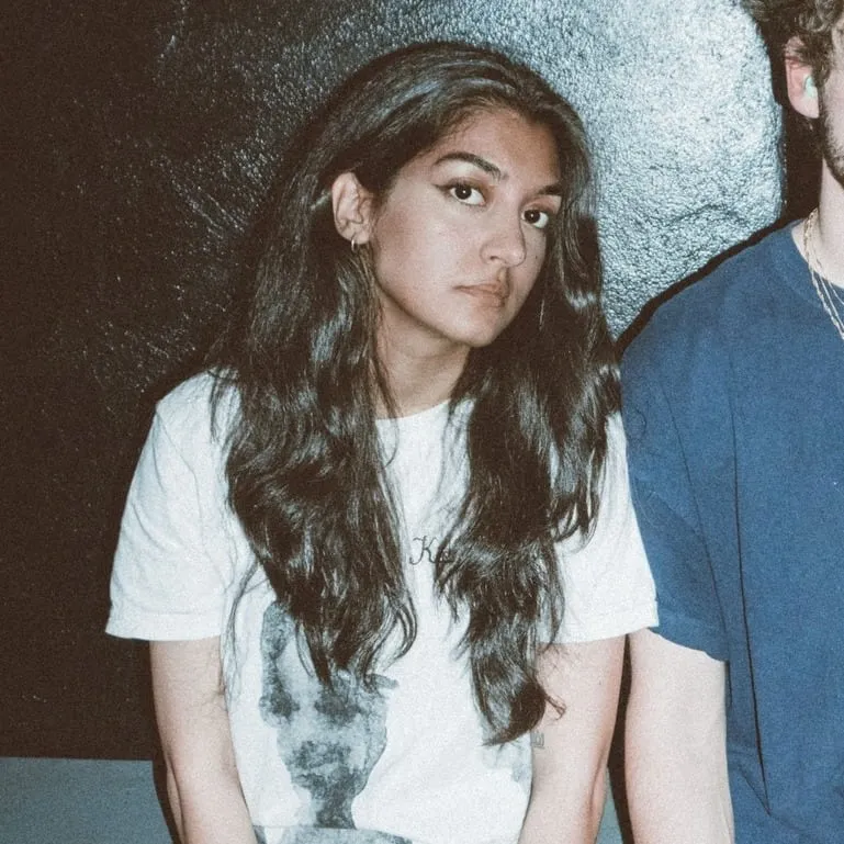 Young woman with long dark hair and neutral expression, sitting against a dark textured background, showcasing modern youth c