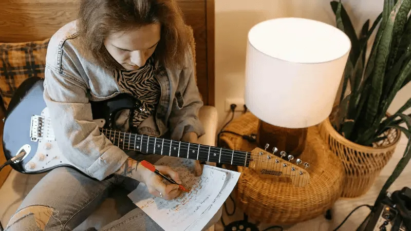 Guitarist writing music notes on sheet music with an electric guitar nearby in a cozy indoor setting.