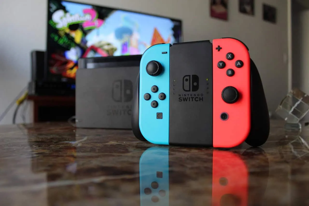 Nintendo Switch console with neon blue and red controllers on a wooden table in front of a TV playing a Nintendo game.