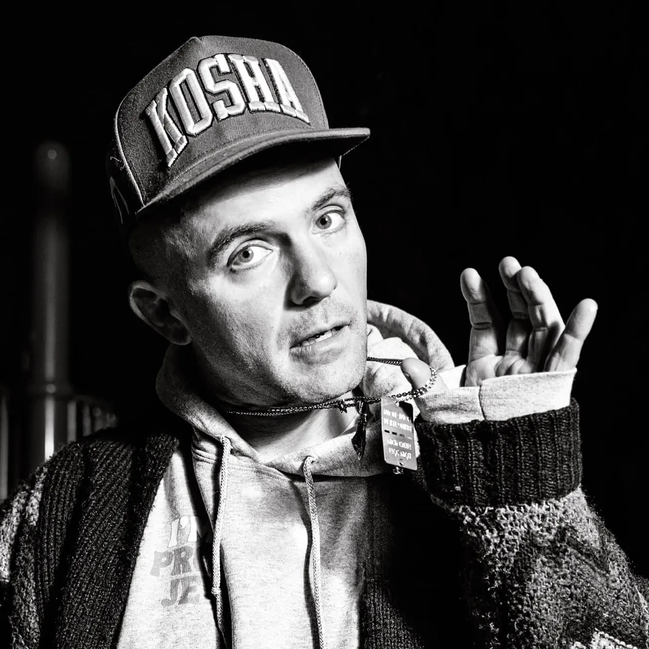 Young man wearing a "KOSH" cap, hoodie, and sweater, making a hand gesture, candid style, black and white photograph, express