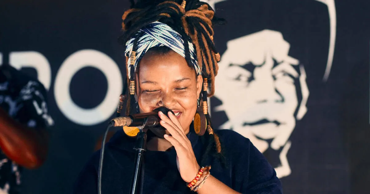 A vibrant female singer with dreadlocks and colorful headwrap performing passionately into a microphone at a music event.