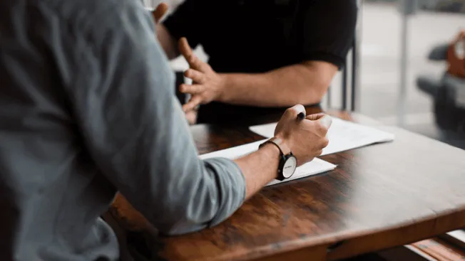 A person signing a document at a wooden table during a business meeting or professional discussion.