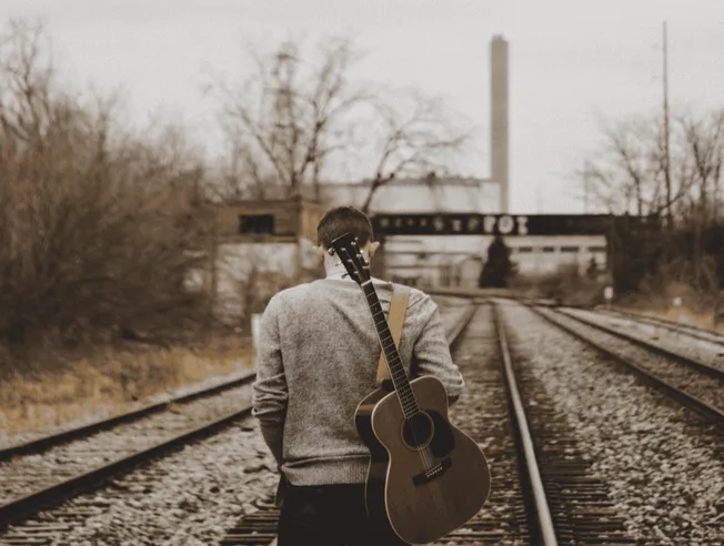 A man walking on railroad tracks with guitar slung over shoulder in a sepia-toned outdoor setting.
