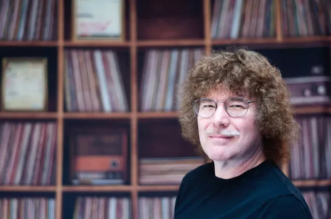Vintage musician with round glasses and curly hair in front of a vinyl record collection on wooden shelves.