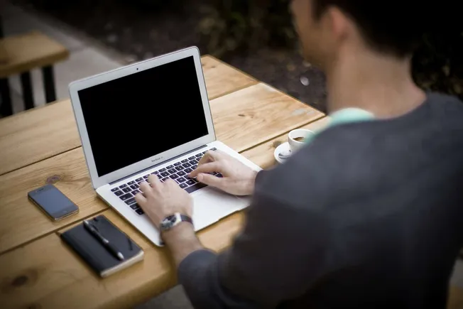 Laptop computer on a wooden table with a smartphone, notebook, pen, and cup of coffee, outdoors setting for remote work or di