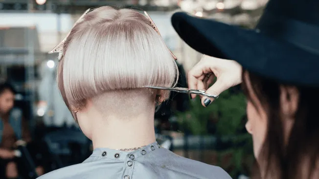Bold hairstylist trimming a model’s short pastel pink bob with an undercut, in a busy salon environment, showcasing professio