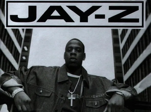 Young Black male artist Jay-Z wearing a chain and jacket, standing confidently outdoors with urban architecture and a large "
