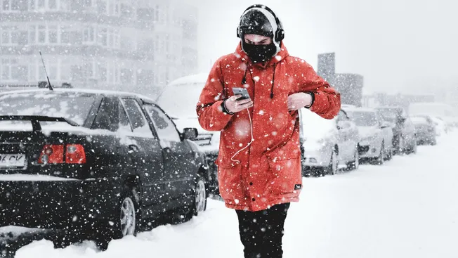 A person in a red winter coat and black face mask walking through a snowy city street while using a smartphone, with parked c
