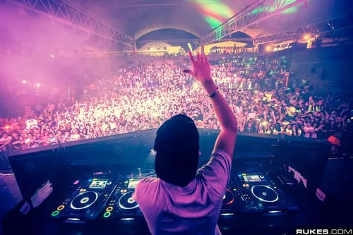 Vibrant electronic dance music concert with a DJ performing on stage amidst a large, energetic crowd under colorful lights.