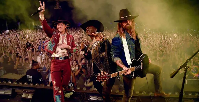 Bright concert performers wearing cowboy hats and colorful outfits on stage with a lively crowd audience in the background ca
