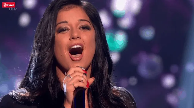 A woman singing passionately into a microphone during a live performance, with a vibrant, bokeh-lit background. The image cap