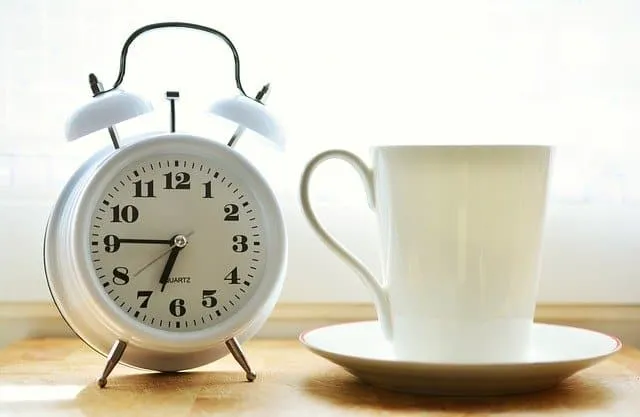 Vintage white alarm clock next to a white coffee cup and saucer on a wooden surface, morning breakfast setting, natural light