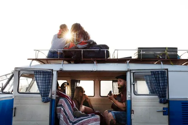 Adventure travelers enjoying a road trip in a classic van with luggage rooftop, outdoor sunlight, and friends creating memora