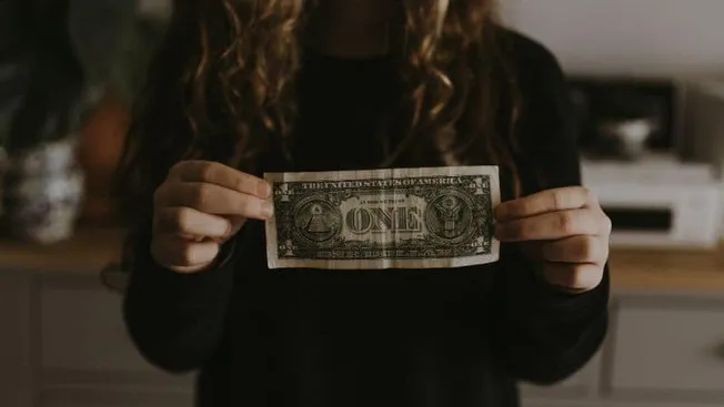 One dollar bill held by a person in a black long-sleeve shirt, small home interior background, symbolizing finance or money c