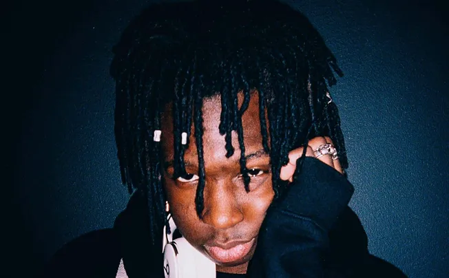 Dreadlocked person with dark skin and rings on fingers, posing against a dark textured background, showcasing urban fashion a