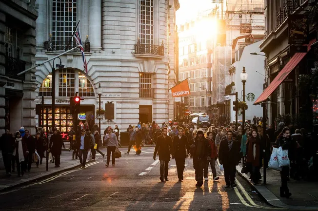 Busy city street in London during sunset, featuring historic architecture, pedestrians crossing, and vibrant urban life.