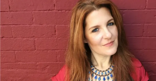 Independent woman with red hair in front of red brick wall, confident and professional, wearing a statement necklace and red