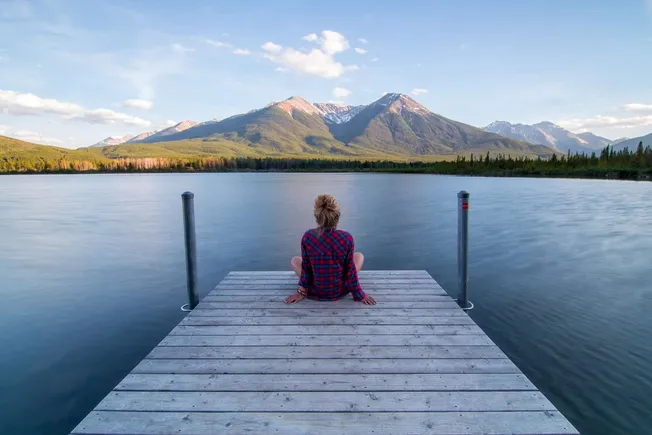 Serene woman sitting on a wooden dock overlooking a tranquil lake with a backdrop of majestic mountains and clear blue sky, c