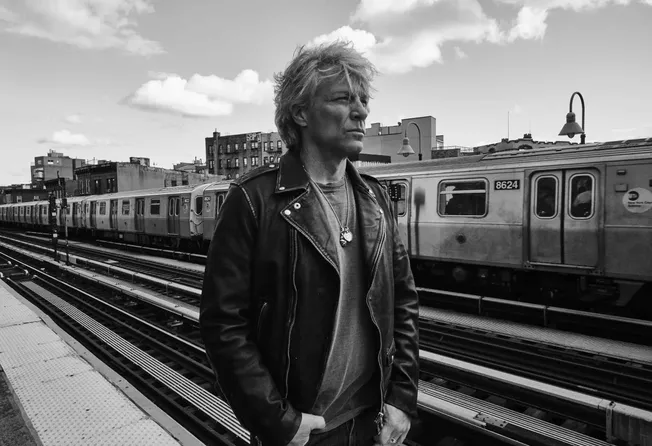 A gritty black-and-white photo of a man with messy hair wearing a leather jacket at a subway station, capturing an urban, edg