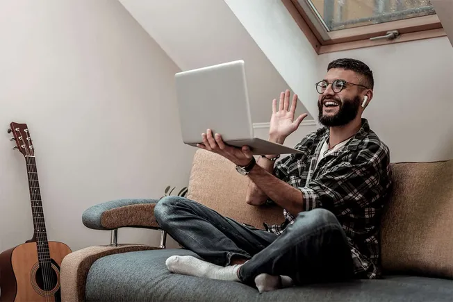 Man engaging in virtual meeting or video call using laptop, wearing wireless earbuds, sitting on sofa in home setting with na