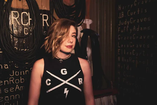 Young woman with short wavy hair wearing a band-inspired sleeveless t-shirt at a music studio or venue, surrounded by audio c
