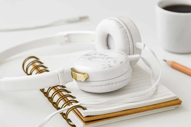 White over-ear headphones resting on a notebook with a gold paperclip, a cup of coffee in the background, and a pencil on a c