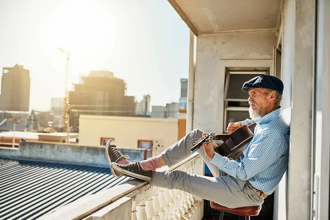 A senior man playing guitar on a balcony at sunset, enjoying music in an urban setting, representing creativity and leisure f