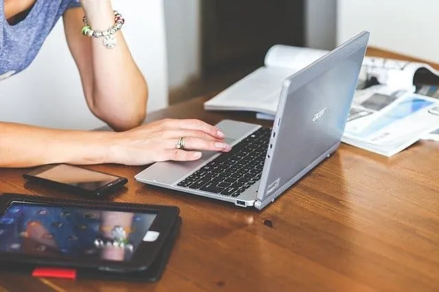 A person working on a silver laptop at a wooden desk with a smartphone and magazines nearby, demonstrating digital marketing