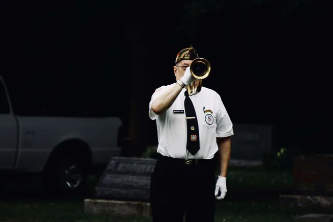 Patriot playing bugle at a memorial ceremony honoring military veterans, wearing ceremonial uniform with badges and patriotic