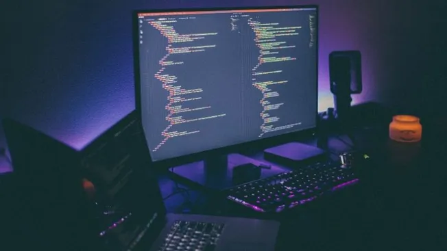 High-tech computer setup with multiple monitors displaying coding scripts, surrounded by LED lighting, speakers, and a keyboa