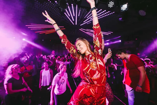 Vibrant woman dancing with eyes closed at a lively nightclub with colorful LED lights and a crowd enjoying music.