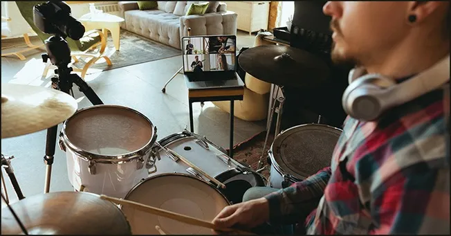 A person practicing drums at home during a virtual band rehearsal, with a laptop displaying remote musicians, set in a cozy l