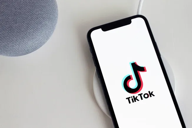 TikTok logo displayed on a smartphone screen next to a smart speaker on a white surface.