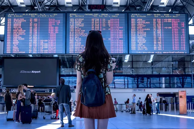 Flights departure and arrival digital display at a busy airport terminal, woman with backpack waiting, travelers with luggage