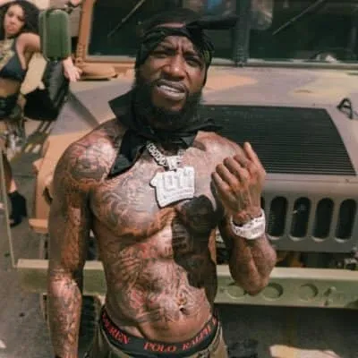 A shirtless man with tattoos, wearing jewelry and a bandana, standing in front of a Jeep during daytime outdoor event.