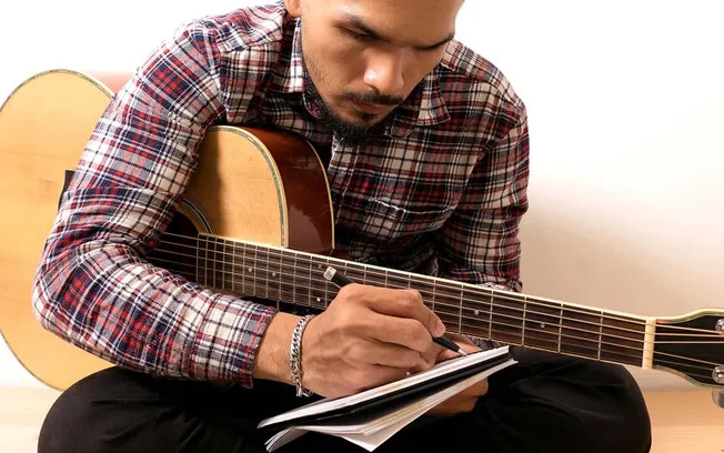 Guitar player writing notes with a pen on a notepad, sitting indoors. Focus on musician engaged in songwriting or practice wi