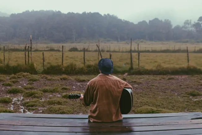 Cityscape, outdoor music performance, person playing acoustic guitar on wooden bench, nature setting with rolling hills, fenc