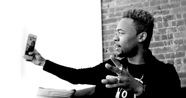 Young man taking a selfie with smartphone, urban setting with brick wall, black and white photo, modern social media, persona