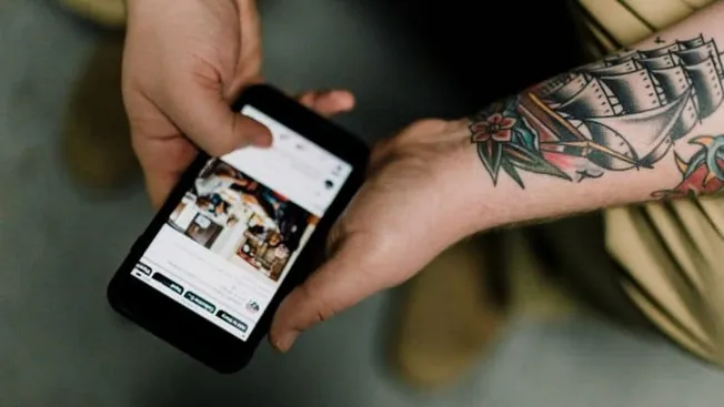 Hand holding smartphone with social media app open, showcasing digital marketing and social networking concepts, tattoos visi