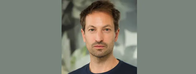 Portrait of a man with brown hair and blue eyes, wearing a black shirt, in an indoor setting with blurred greenery in the bac