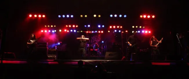 Guitarists and drummer performing live on stage with vibrant red, blue, and yellow stage lighting, showcasing a dynamic conce