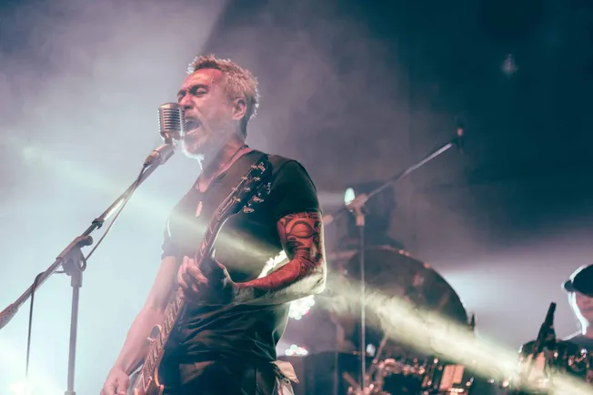 Vocalist guitarist performing live at a music concert, with intense expression and tattoos, illuminated by stage lights, emph