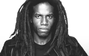 A young man with long dreadlocks and intense expression, wearing a black leather jacket, against a plain background.