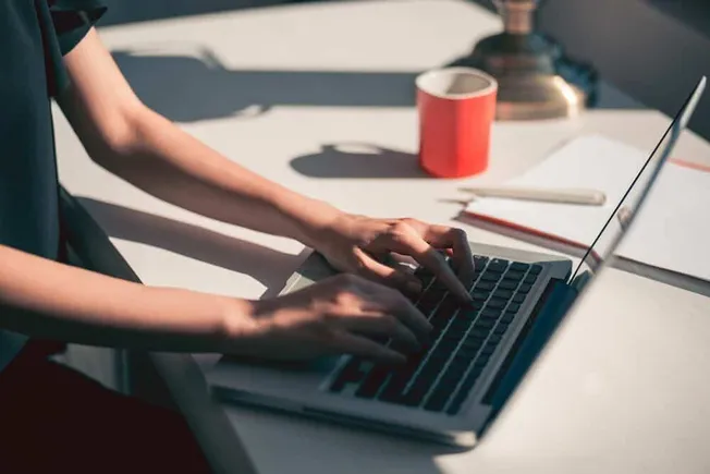 Woman typing on a laptop at a work desk with stationery and a lamp in the background.