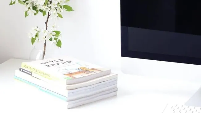 Stacks of fashion and branding magazines on a white desk with a minimalist floral arrangement and a computer monitor, illustr