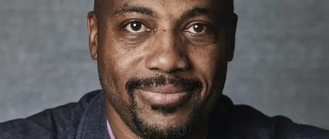 Celebrity portrait of a man with a beard and mustache, smiling, against a gray background.