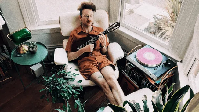 Man playing guitar in cozy home setting with vinyl turntable and green plants.
