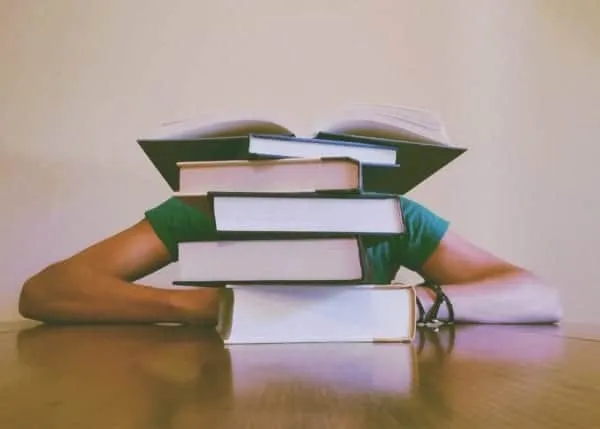 Cluttered desk with stacked books and a person hidden behind, representing study overload or information chaos.