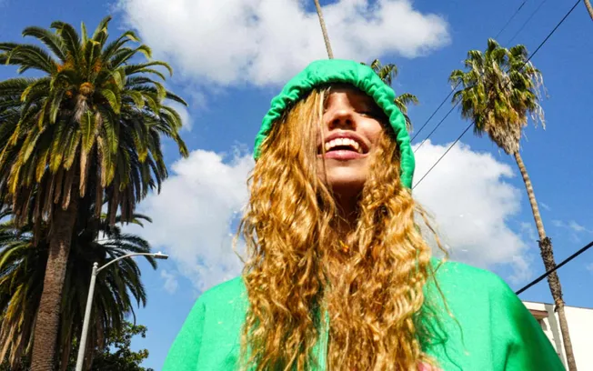 Bright young woman with long, curly red hair wearing a vibrant green hoodie outdoors amid palm trees and blue sky, showcasing