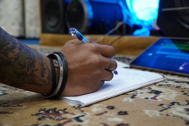 Hand writing in a notebook with a pen, tattoos visible on the wrist, music equipment in the background, casual creative works